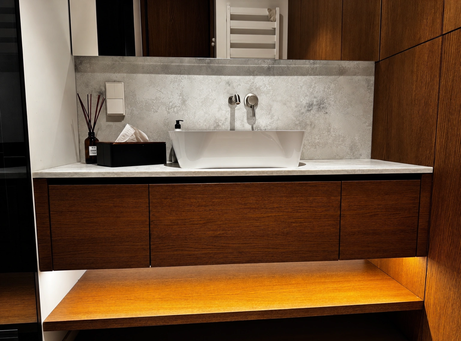 A white washbasin on a countertop, with brown cabinets and an illuminated shelf beneath it.