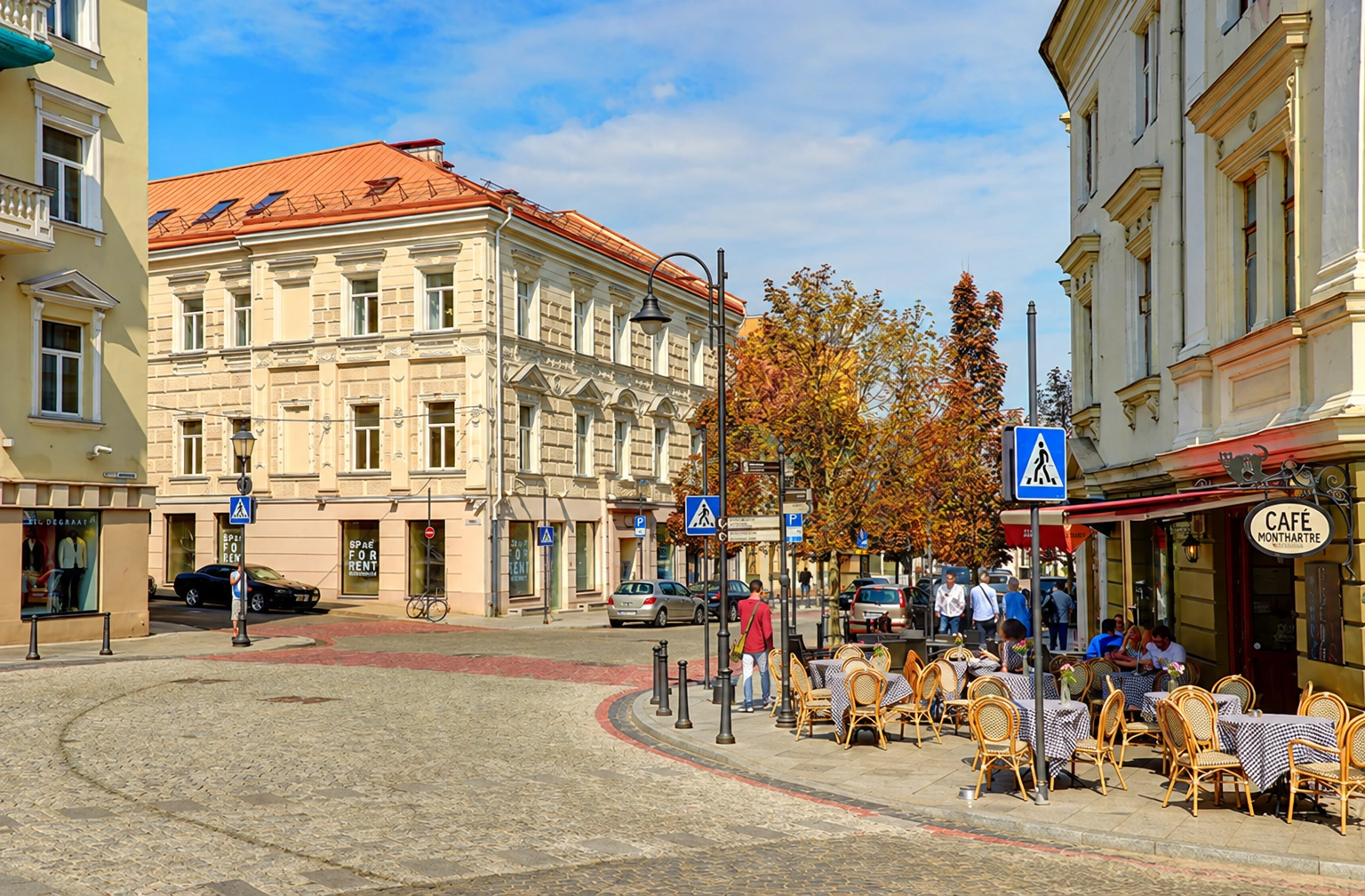 Gatvė tarp daugiaaukščių pastatų, dešinėje – lauko kavinės staliukai ir kėdės.