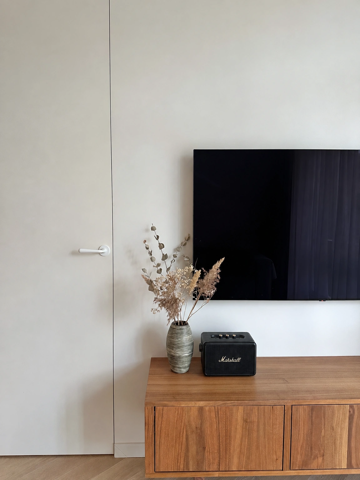 An der Wand hängt ein Fernseher, und auf der Kommode stehen eine Vase mit getrockneten Pflanzen und ein Lautsprecher.