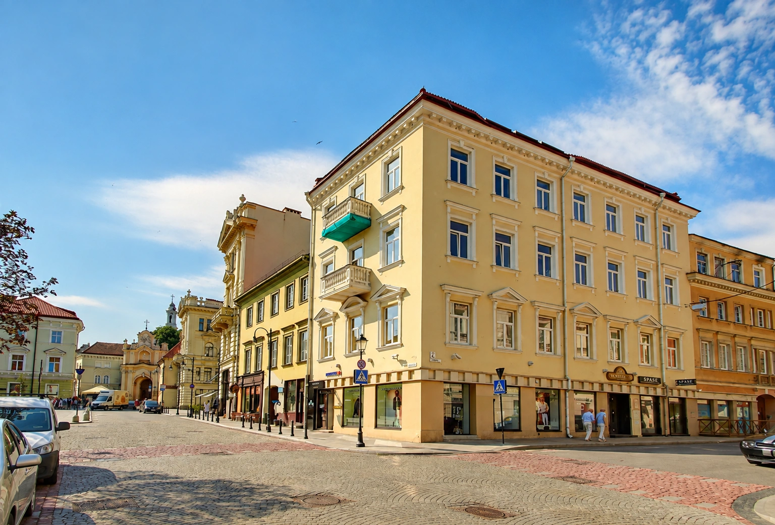 Geltonas kampinis pastatas su balkonais ir vitrininiais pirmo aukšto langais prie gatvių sankryžos.