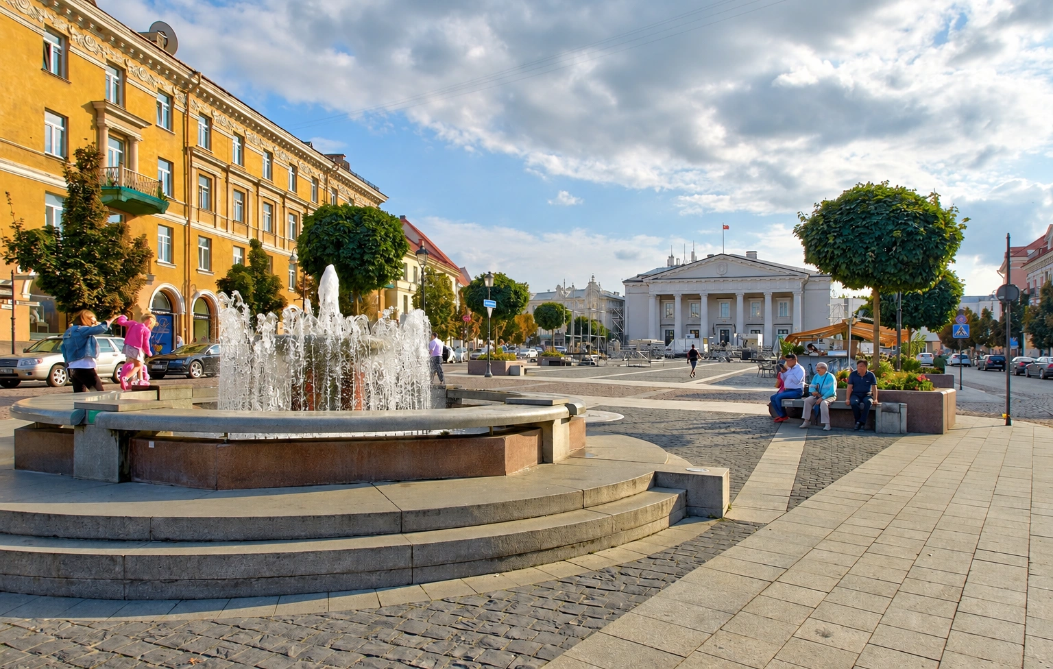 A city square with a fountain and a yellow multi-storey building nearby.