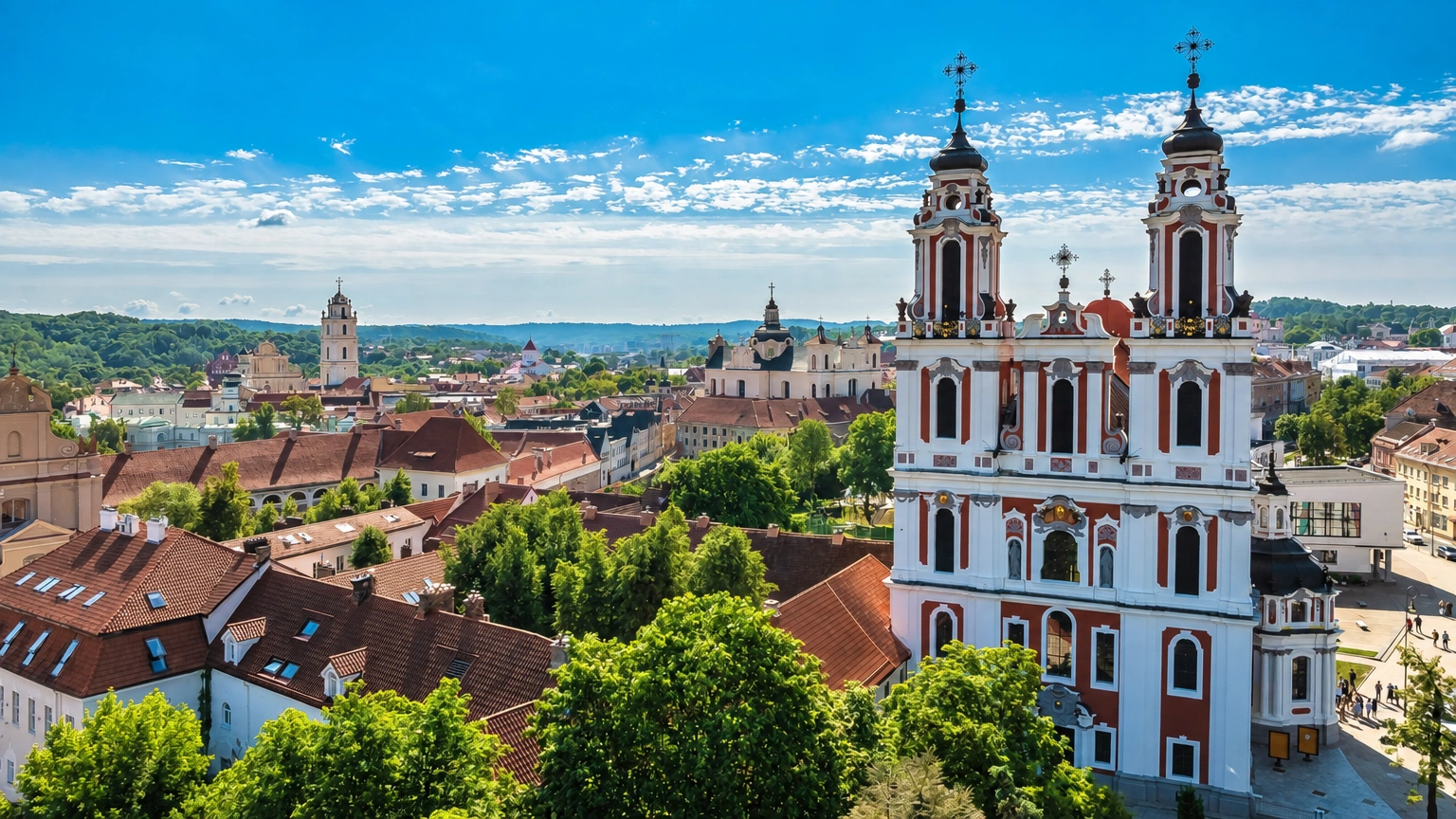 Ein Stadtpanorama mit einer zweitürmigen Kirche, roten Gebäudedächern und grünen Bäumen.