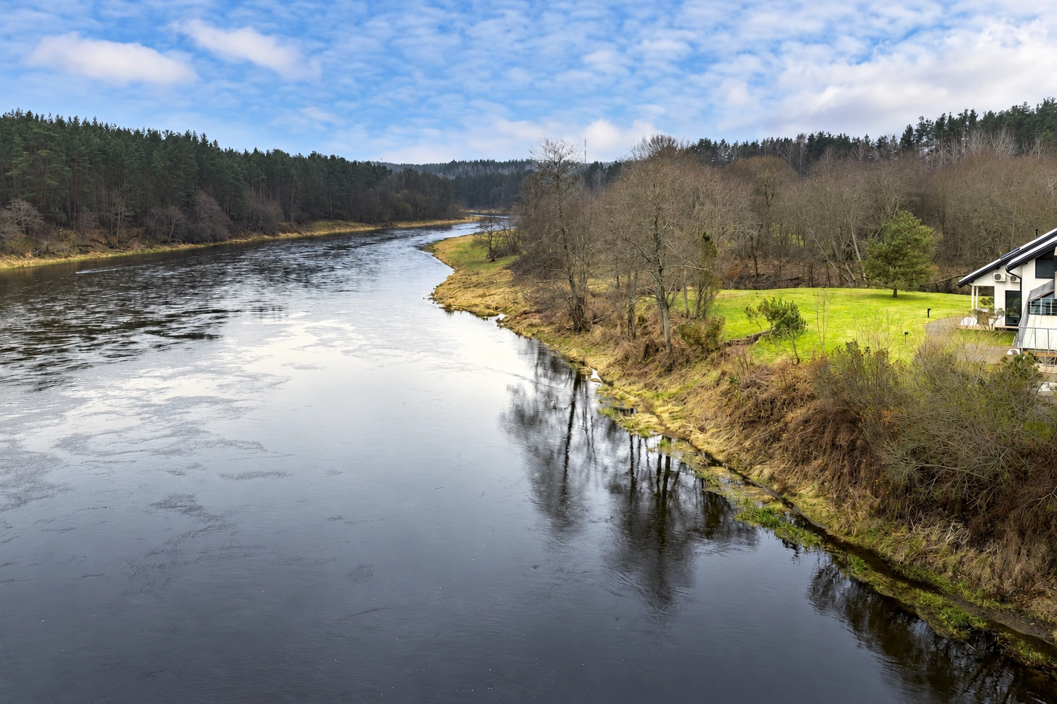 Ein Fluss fließt zwischen baumbewachsenen Ufern; am rechten Ufer ist ein Teil eines Hauses zu sehen.