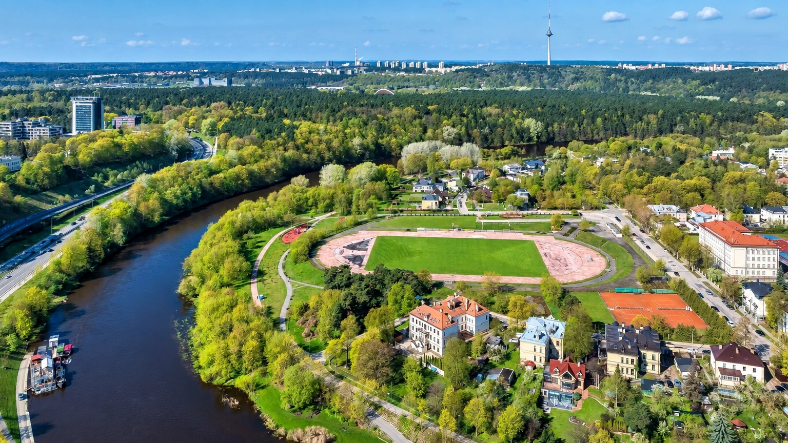 From above, a river, a stadium with a running track, and buildings surrounded by trees can be seen.