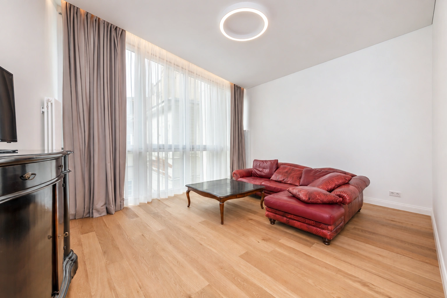 Svetainėje stovi raudona sofa ir stiklinis kavos staliukas, didelius langus dengia užuolaidos.