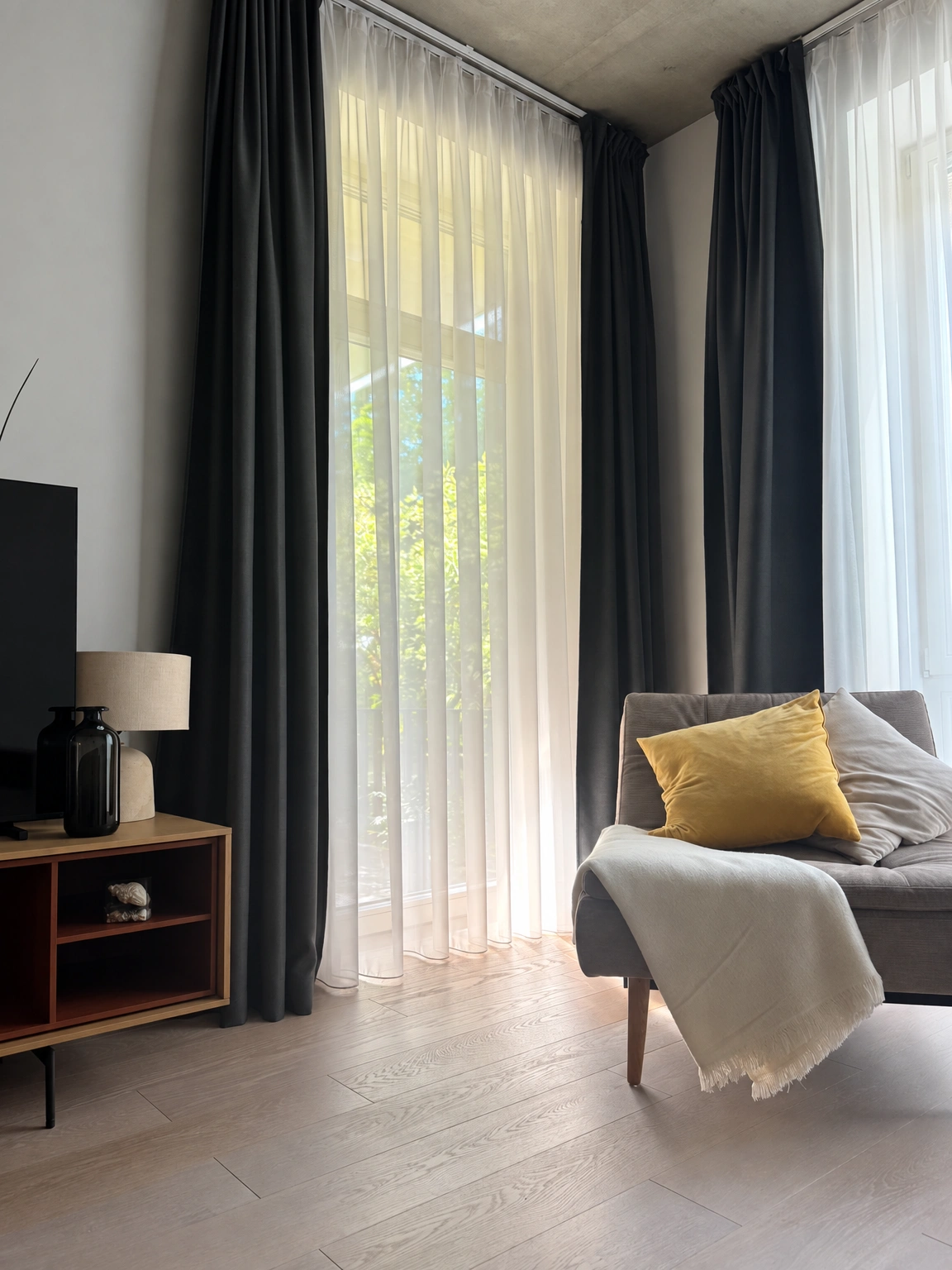 Prie didelių langų su tamsiomis užuolaidomis stovi pilka sofa su geltona pagalvėle.