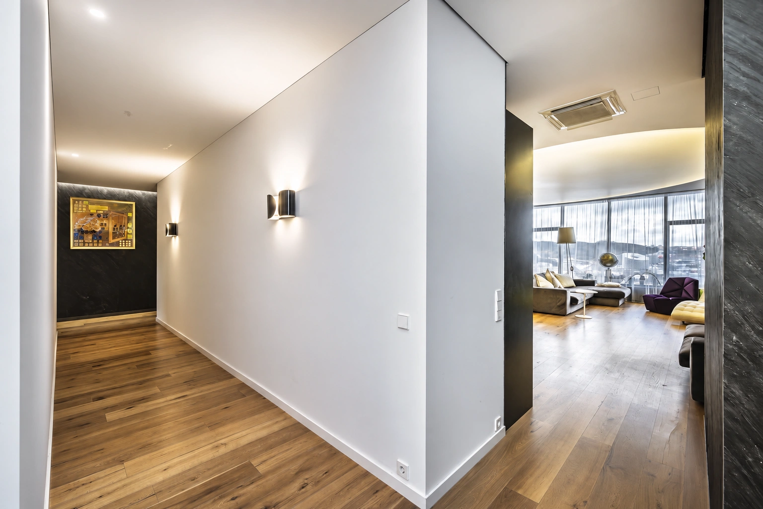 A corridor with white walls; on the right, a living room with large windows is visible through an open passageway.
