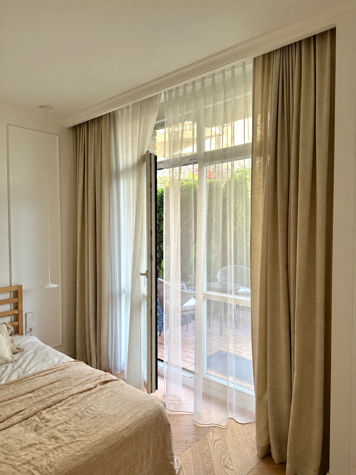 Im Schlafzimmer sind ein Bett und hohe Fenster mit transparenten und bräunlichen Vorhängen zu sehen.