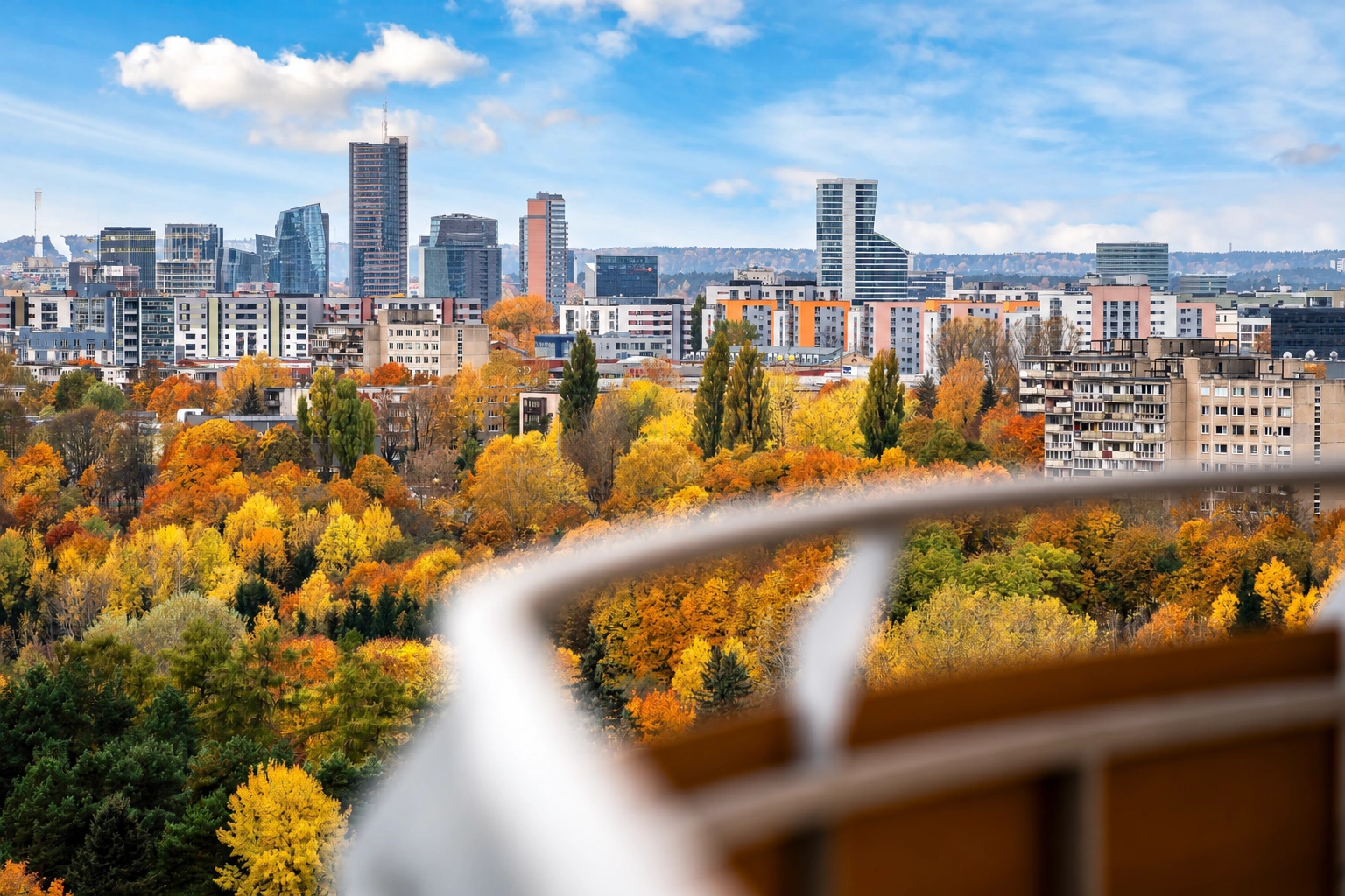 Stadtpanorama mit hohen Gebäuden und herbstlichen Bäumen; im Vordergrund ist ein Balkongeländer zu sehen.