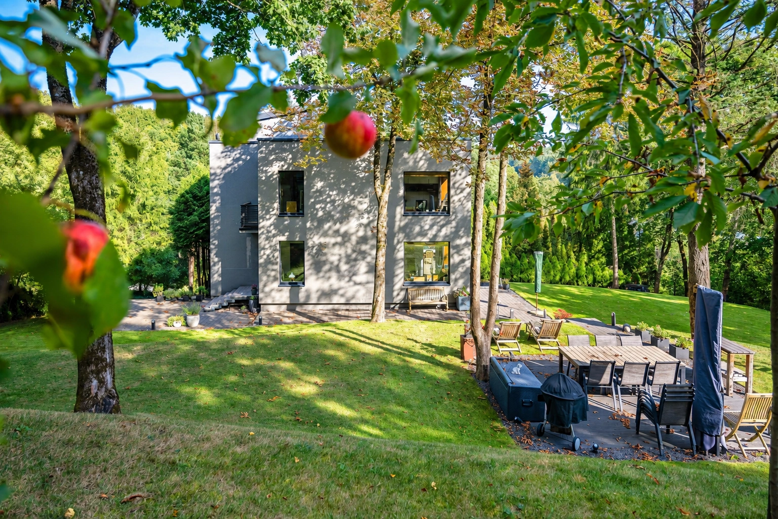 Ein von Bäumen umgebenes Haus; daneben befinden sich eine Rasenfläche und eine Terrasse mit einem Tisch und Stühlen.