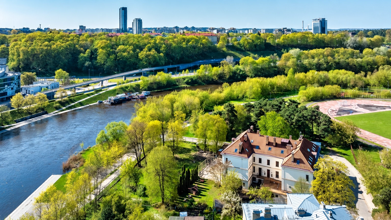 An aerial view shows a building with a pitched roof, trees growing around it, and a river flowing nearby.
