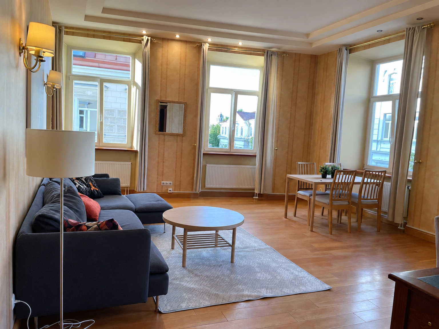 Wohnzimmer mit einem dunklen Sofa, einem runden Couchtisch und einem Esstisch mit Stühlen an den Fenstern.