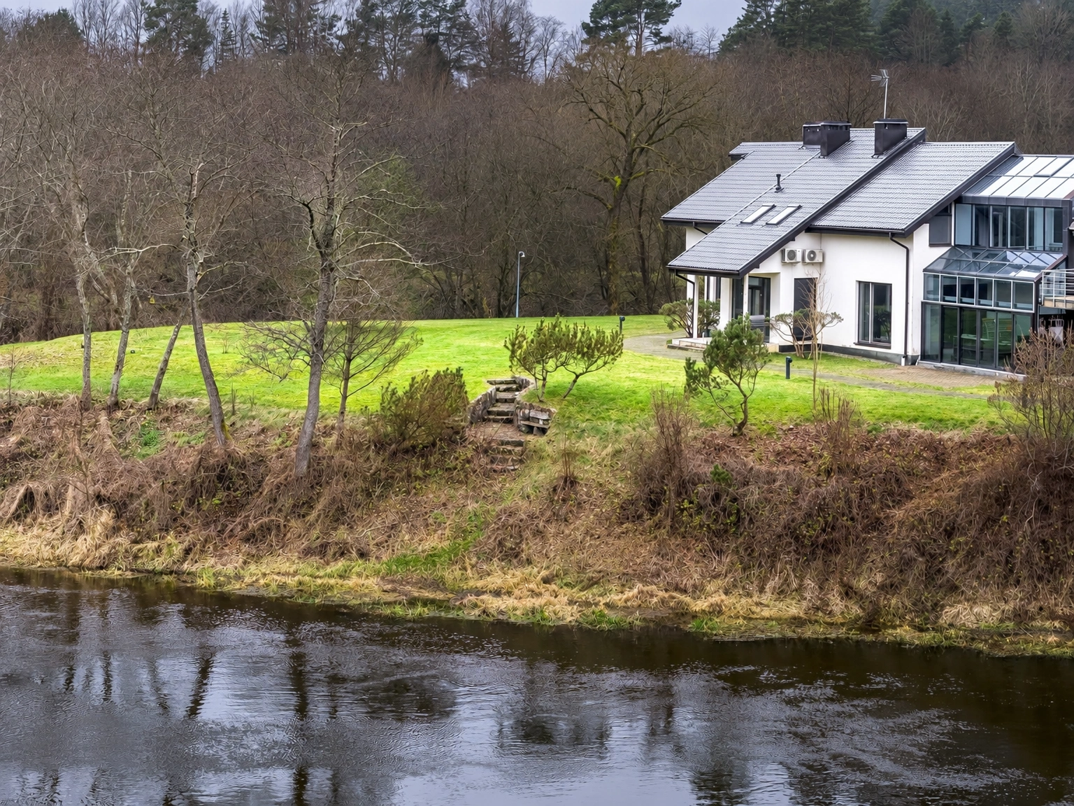 An einem Gewässer steht ein Haus mit großen Fenstern, und im Hof grünt der Rasen.