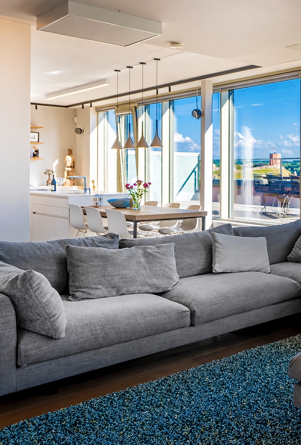 Im Wohnzimmer steht ein graues Sofa mit Kissen; dahinter sind ein Esstisch und große Fenster zu sehen.