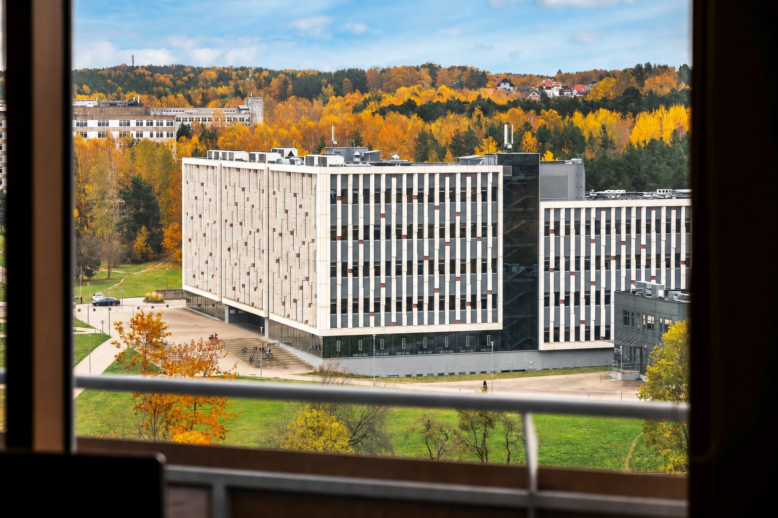 Durch das Fenster sind mehrstöckige Gebäude und herbstliche Bäume zu sehen, im Vordergrund ein Balkongeländer.