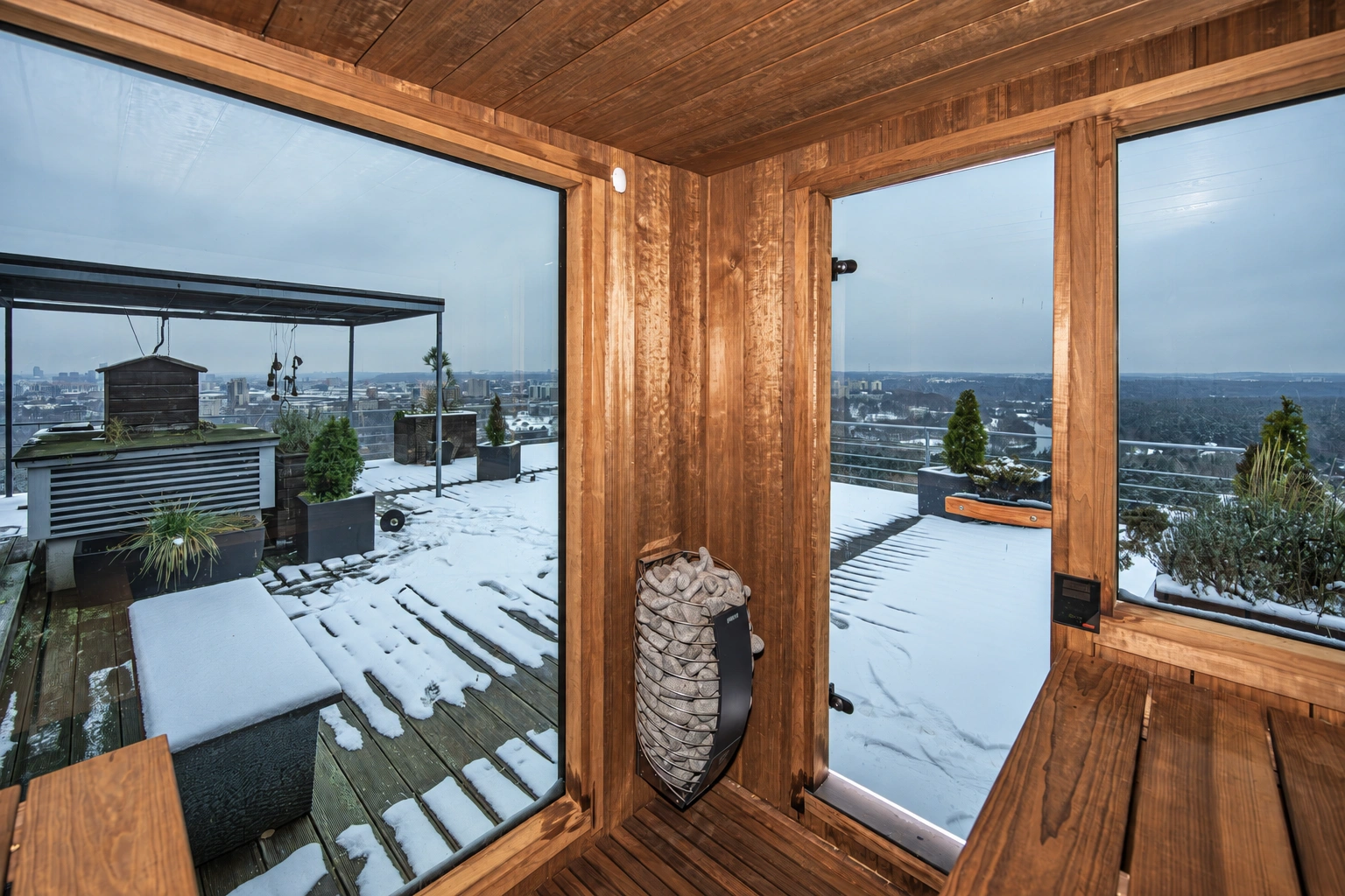 Im Inneren der Sauna sind Bänke, ein Ofen mit Steinen und große Fenster mit Blick auf eine schneebedeckte Terrasse zu sehen.