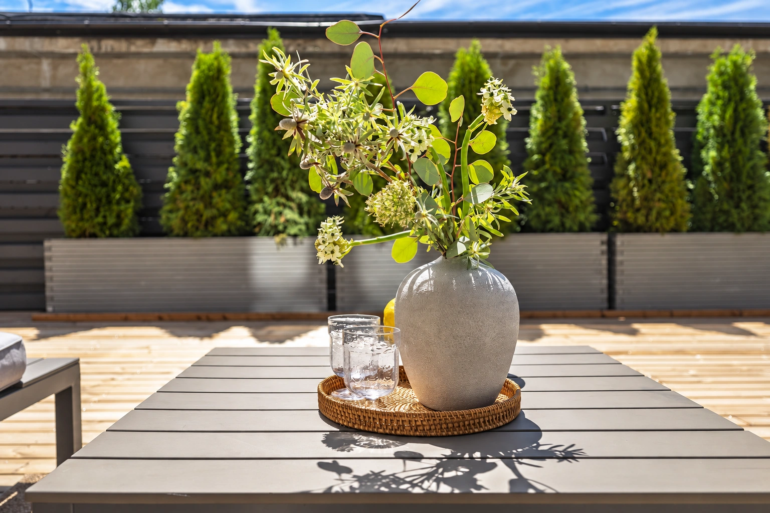 Auf der Terrasse steht ein Tisch mit einer Blumenvase, und entlang des Zauns wachsen Thujen.
