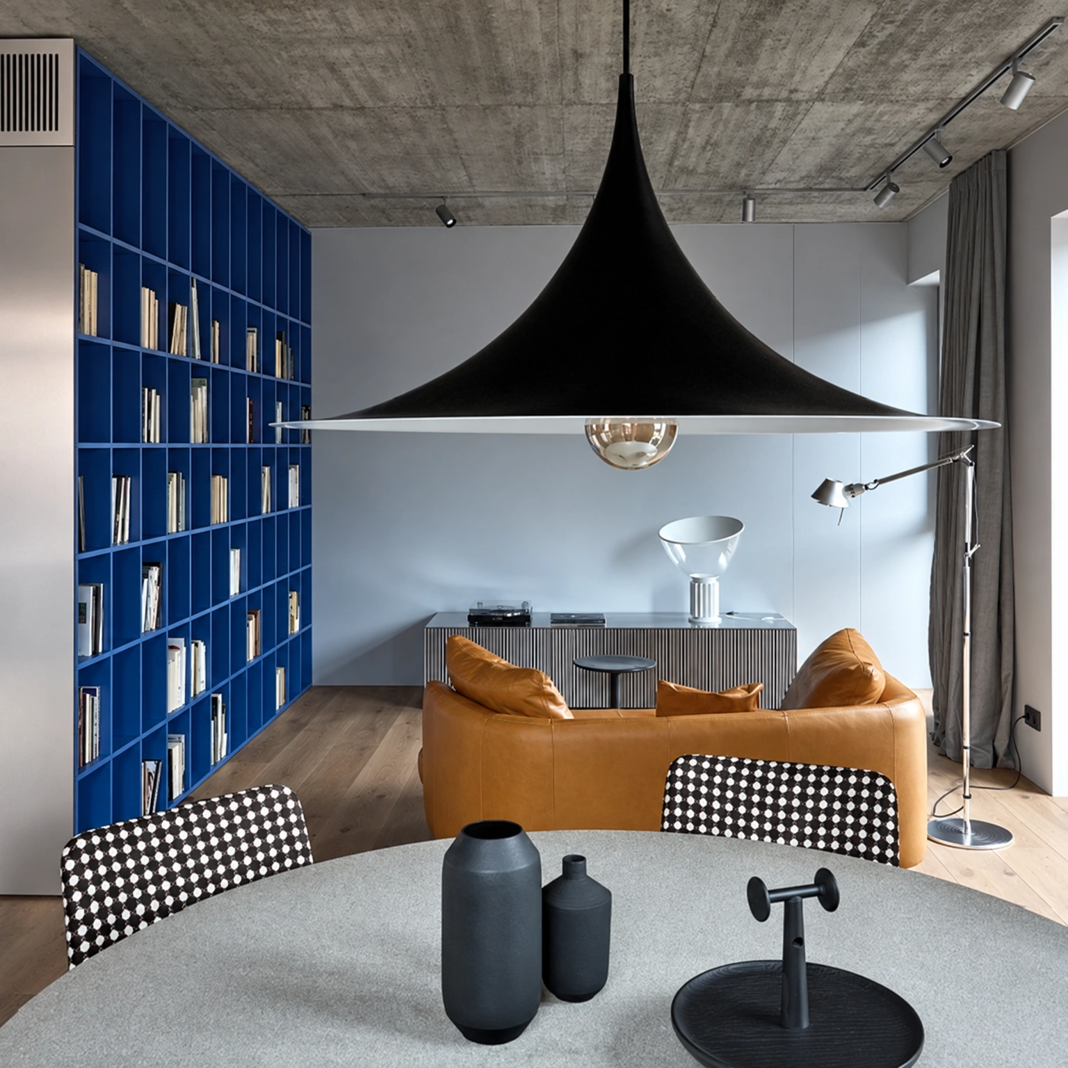 Virš apvalaus stalo su kėdėmis kabo didelis juodas šviestuvas, už stalo matyti sofa.