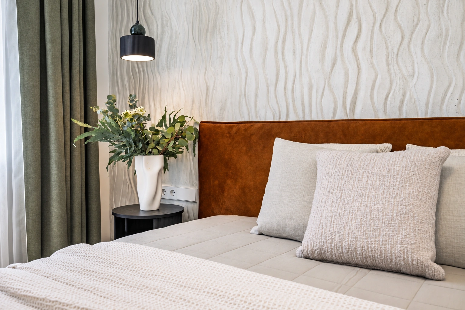 Ein Bett mit Kissen und einem braunen Kopfteil; daneben steht ein kleiner Tisch mit einer Vase mit Blumen.