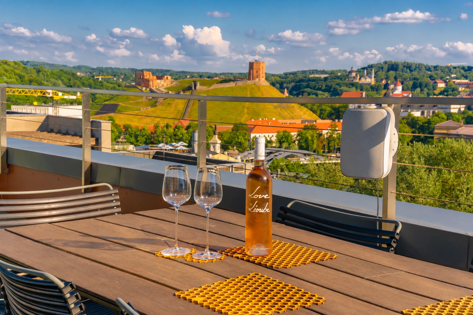 Auf dem Terrassentisch stehen eine Flasche und zwei Gläser; hinter dem Geländer sind grüne Hügel zu sehen.