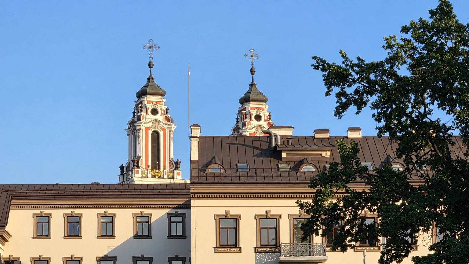 A light-colored building with two towers, its façade partly obscured by the branches of a tree.