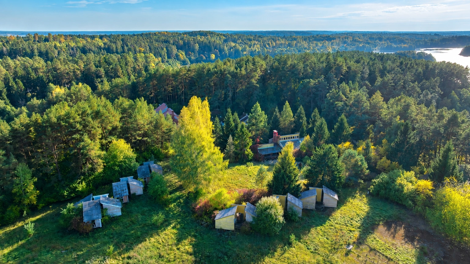 Auf einer von Wald umgebenen Wiese stehen kleine Bauten, und in der Ferne ist ein Gewässer zu sehen.