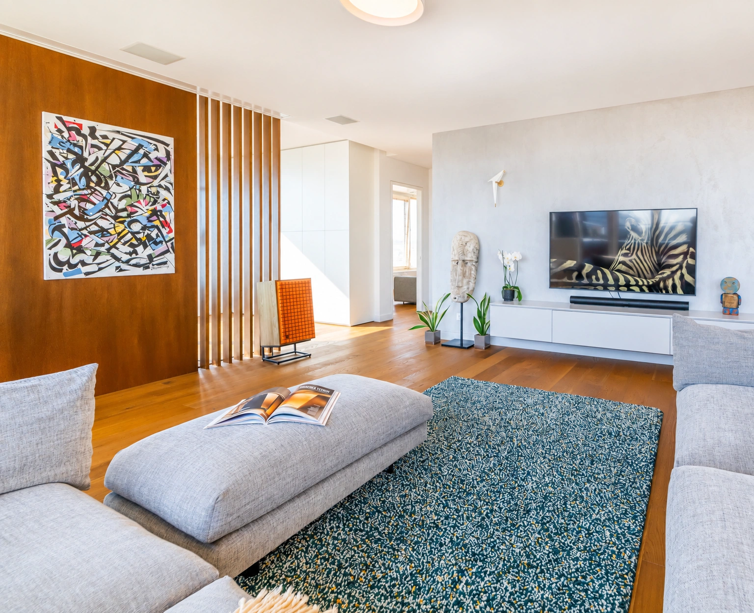Wohnzimmer mit grauen Sofas, einem Teppich und einem an der Wand angebrachten Fernseher.